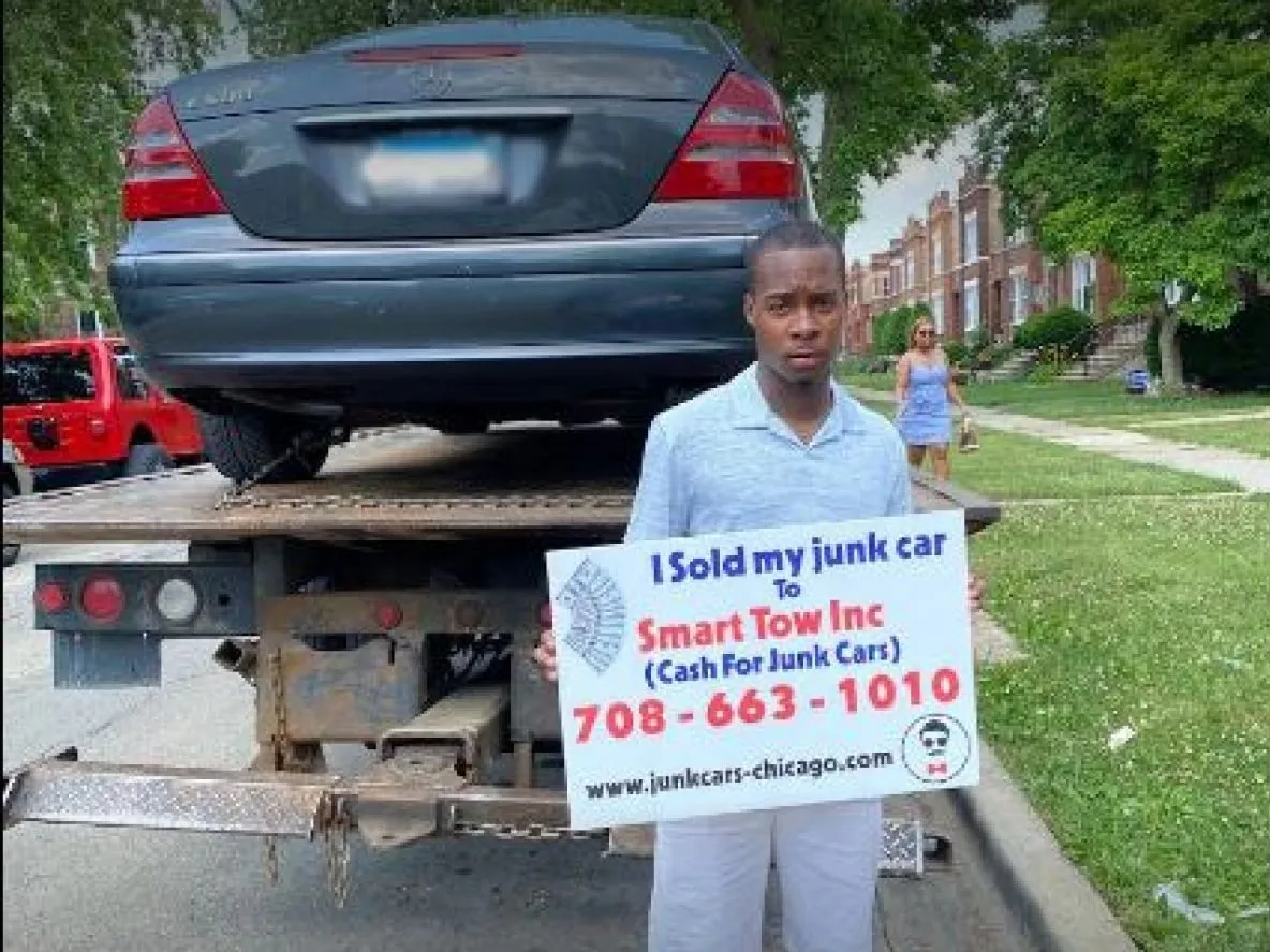 A customer holding a sign that reads I sold my junk car to Smart Tow Inc, with his dark gray Mercedes loaded on a flatbed behind him
